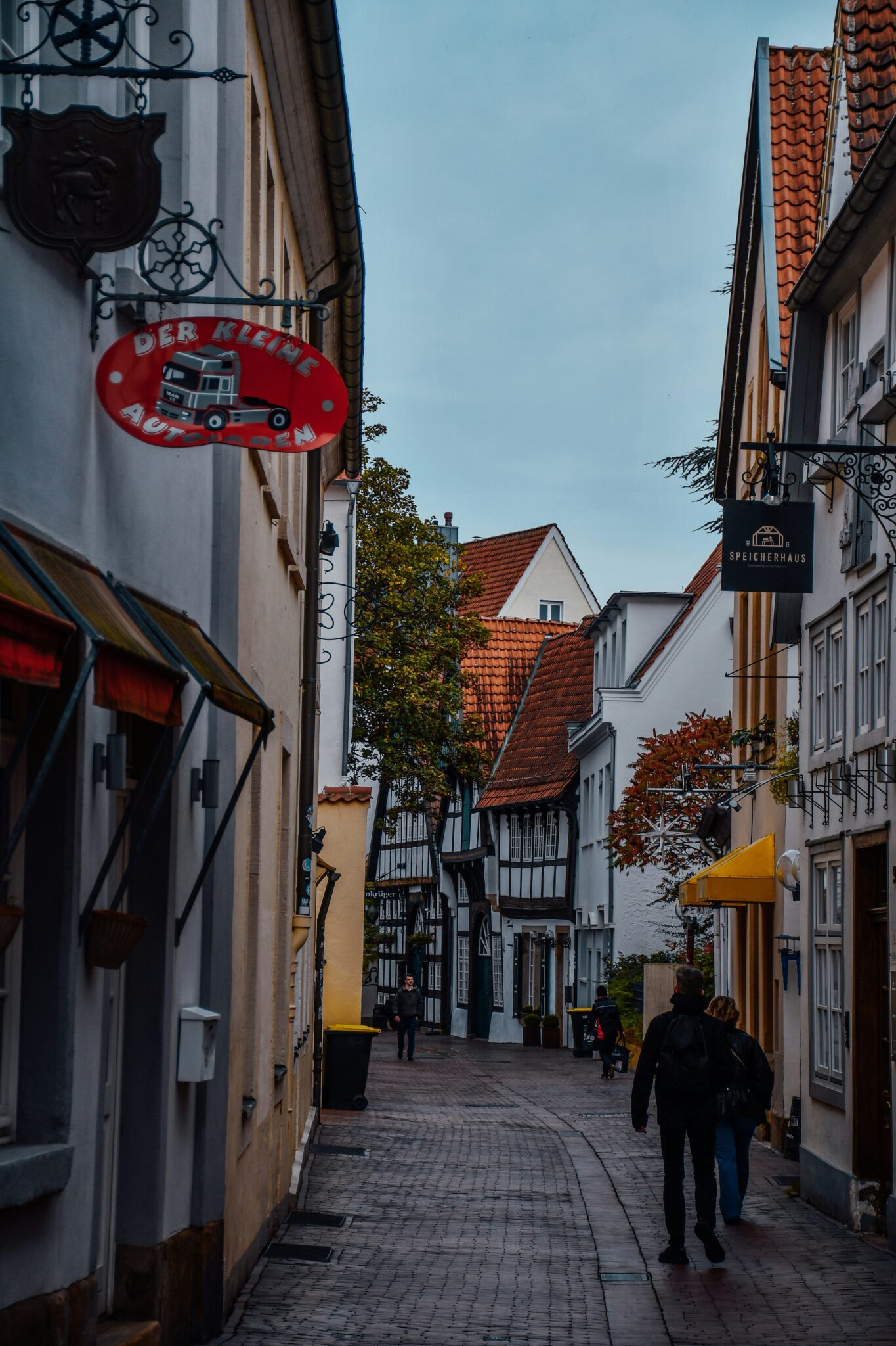 Enge Altstadtgasse mit Fachwerkhäusern, rotem Schild „Der kleine Autoladen“ und Passanten.