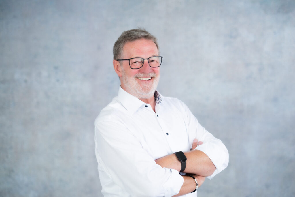 Eine lächelnde Person mit Brille und weißem Hemd vor neutralem grauen Hintergrund.