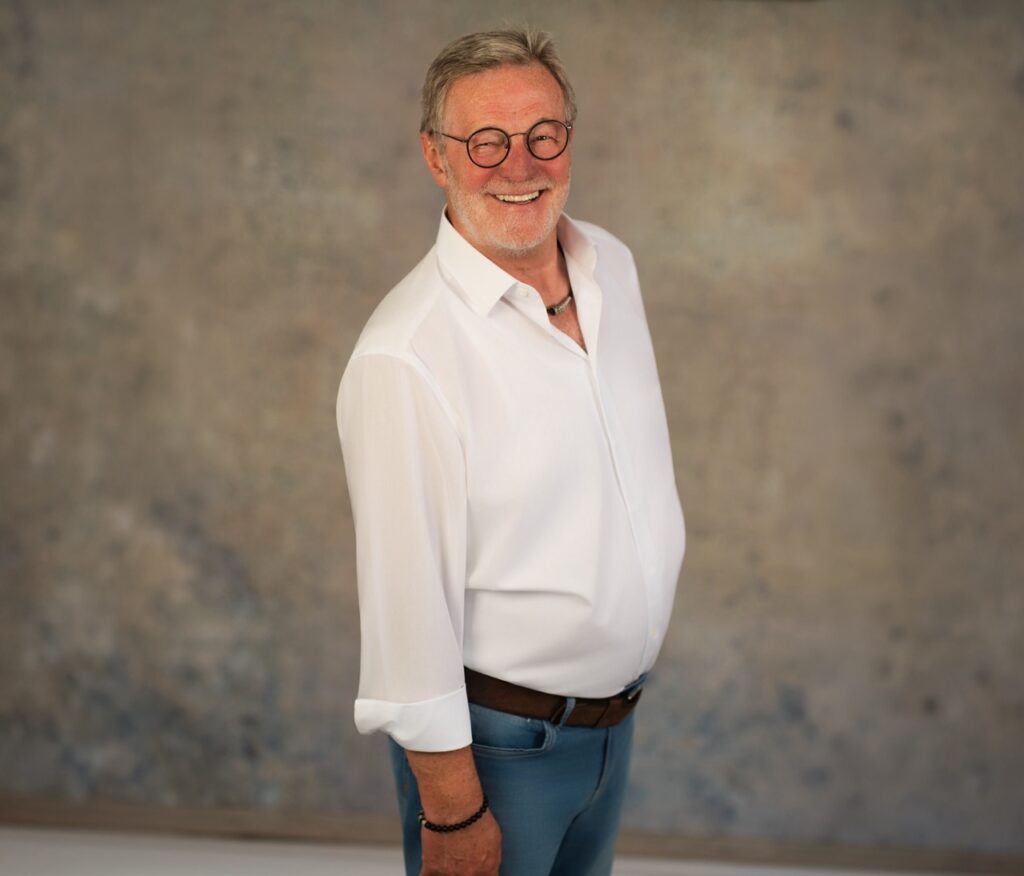 Eine lächelnde Person im weißen Hemd und blauer Hose vor neutralem Hintergrund, freundlich und einladend.