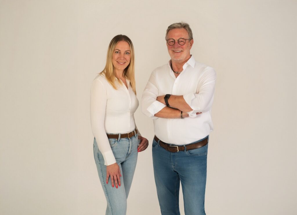 Ein Mann und eine Frau in weißen Hemden und blauen Jeans lächeln entspannt vor hellem Hintergrund.